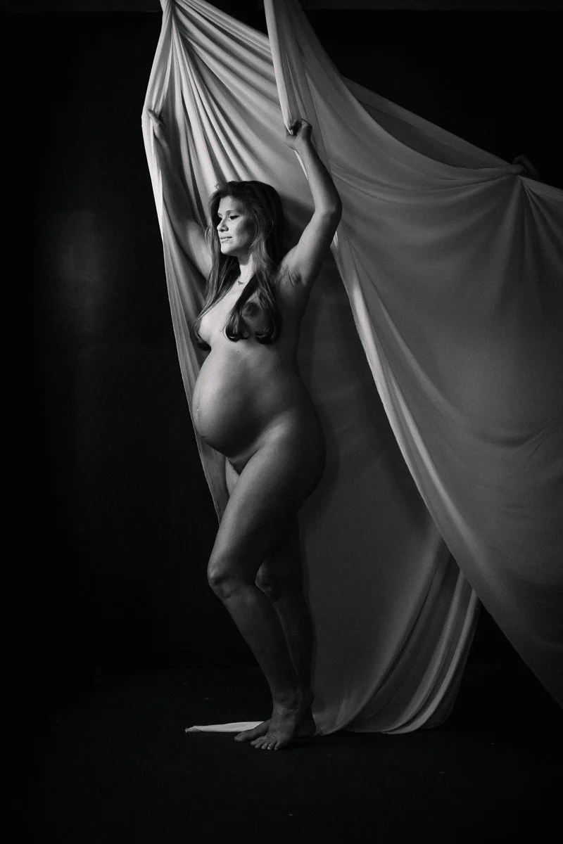A striking black and white fine art maternity portrait of a pregnant woman with arms raised holding flowing fabric, photographed by Emily Scott in Beaufort, SC.