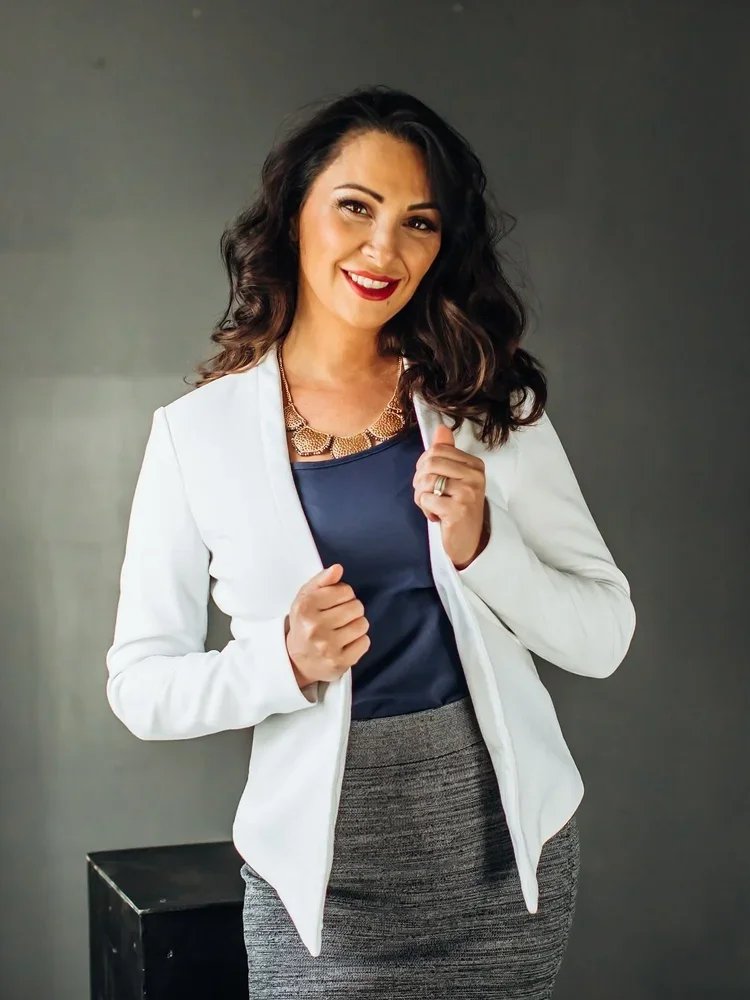 A woman with dark, curly hair smiling, wearing a white blazer over a blue top, a gold necklace, and a gray skirt, standing against a gray background.