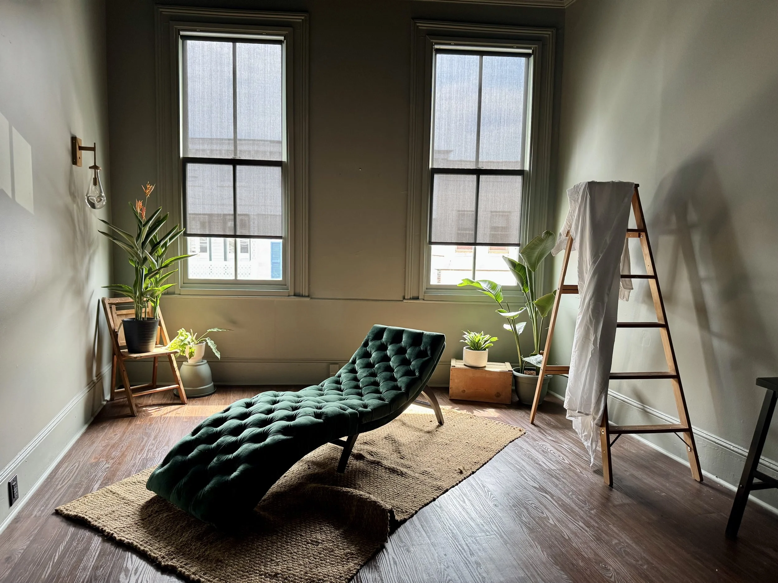 A cozy room with two large windows with blinds, a green tufted chaise lounge on a beige rug, several potted plants, a wooden ladder with a white cloth, and some chairs, all illuminated by natural light.