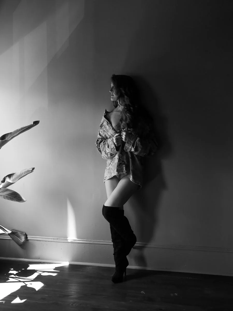A striking black and white fine art boudoir portrait of a woman in an unbuttoned military jacket standing against a studio wall, photographed by Emily Scott in Beaufort, South Carolina.