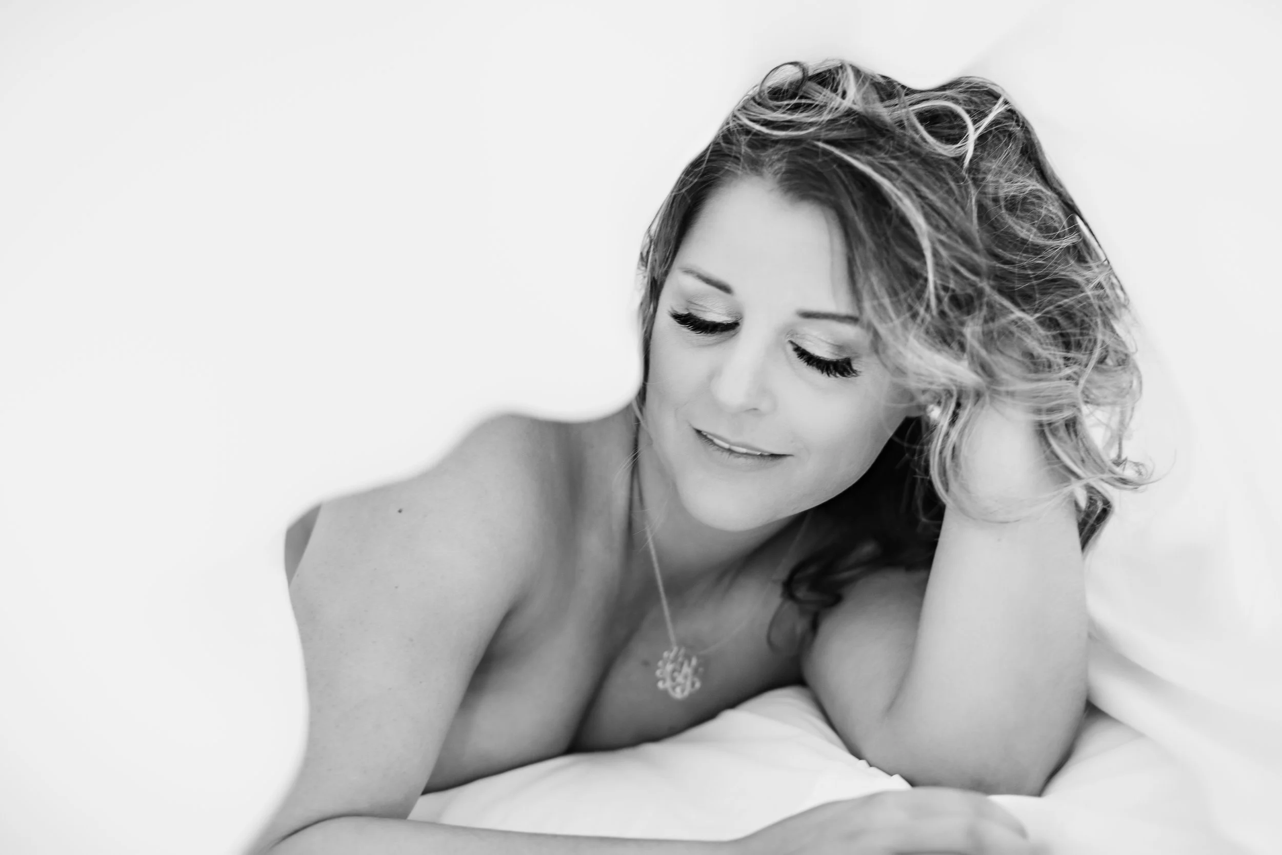 Black and white photograph of a woman with curly hair, lying on a bed, smiling softly with her eyes closed, leaning on her right hand, wearing a necklace, and topless.