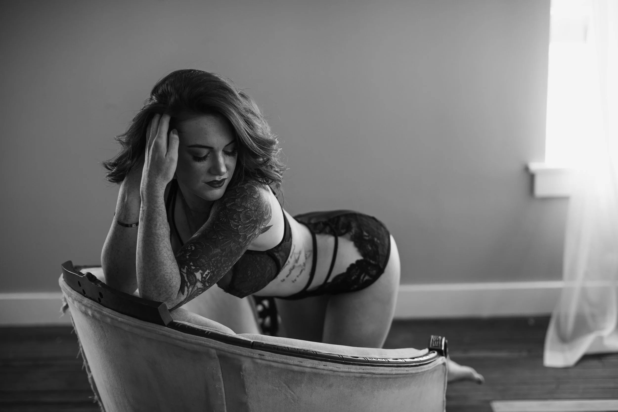 A woman with tattoos, wearing black lace lingerie, posed on her hands and knees on a vintage chair in a softly lit room.