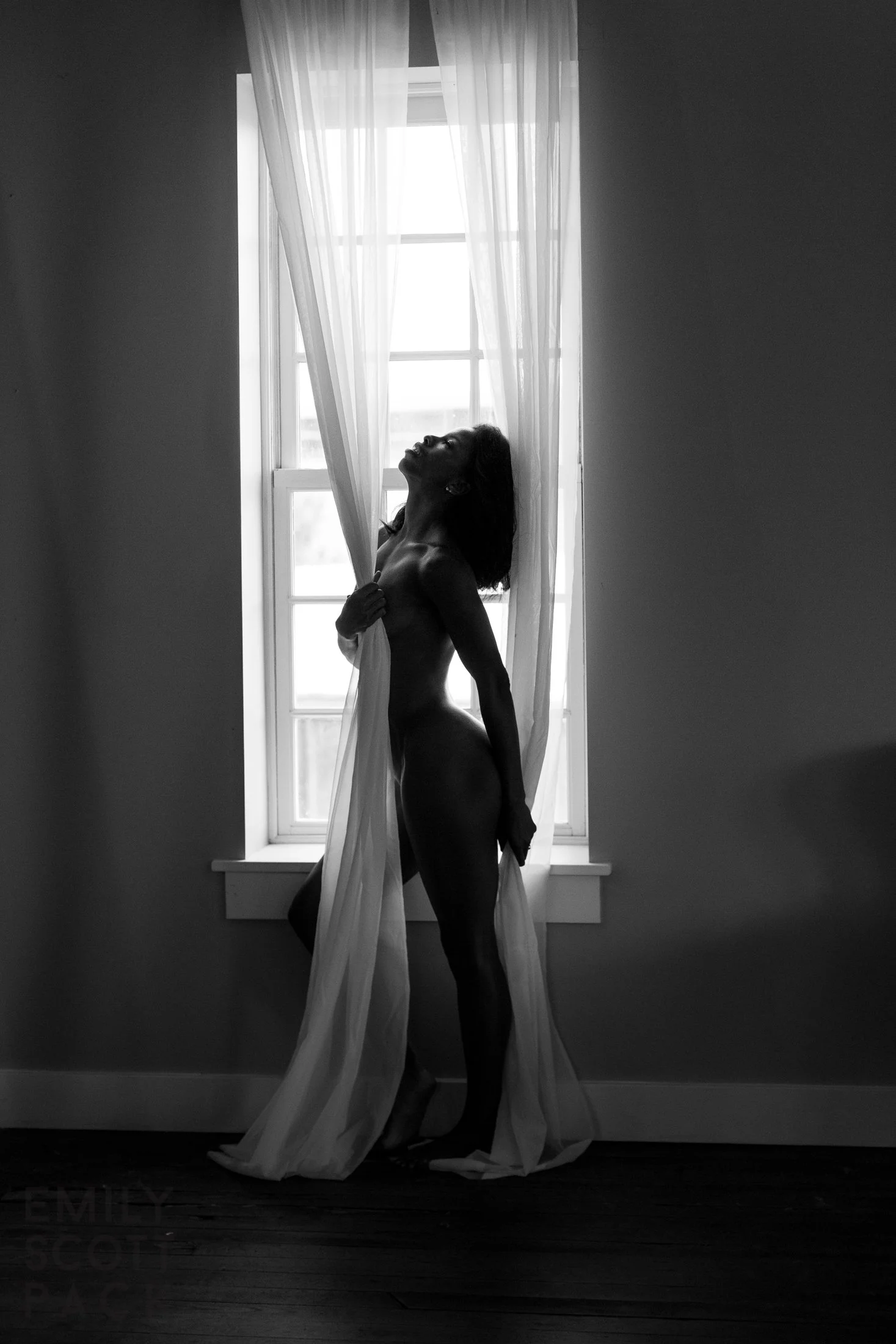 A black and white photo of a woman standing by a window covered with sheer curtains, with light streaming in behind her. She is topless and has her head tilted back with eyes closed, holding the curtain with one hand.
