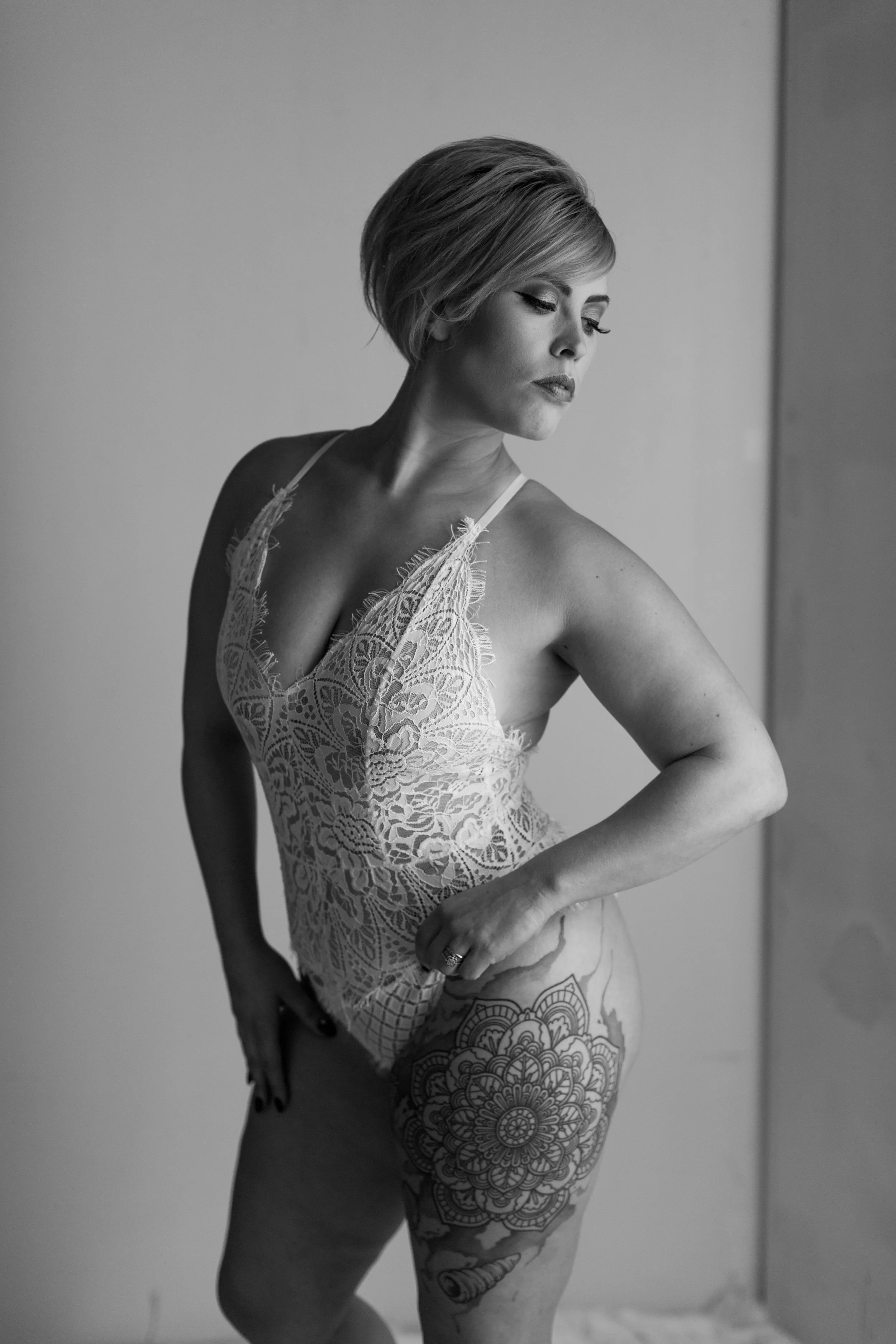 A woman with short hair posing in front of a plain wall, wearing lace lingerie and showcasing a floral tattoo on her thigh in black and white.