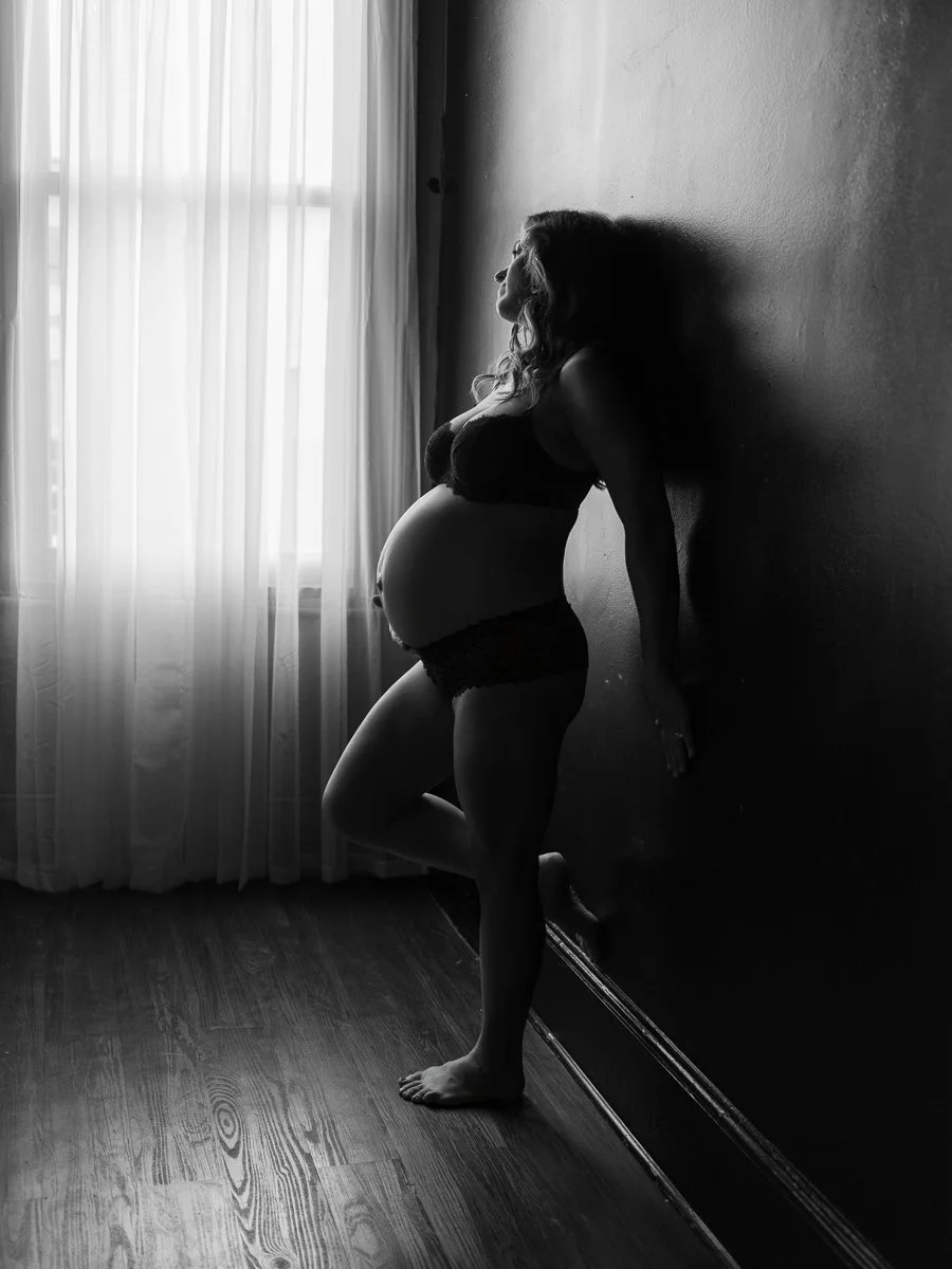 A dramatic black and white maternity boudoir portrait of a pregnant woman leaning against a wall in natural window light, photographed by Emily Scott in Beaufort, SC.