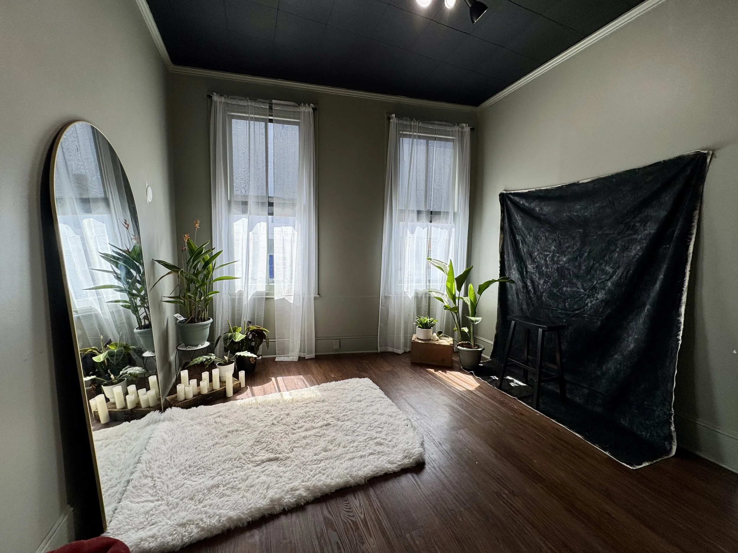 A cozy room with three windows covered by sheer white curtains, hardwood floors, a full-length mirror on the left, a fluffy white rug on the floor, potted plants near the window, and a black backdrop with a small wooden stool in the corner.