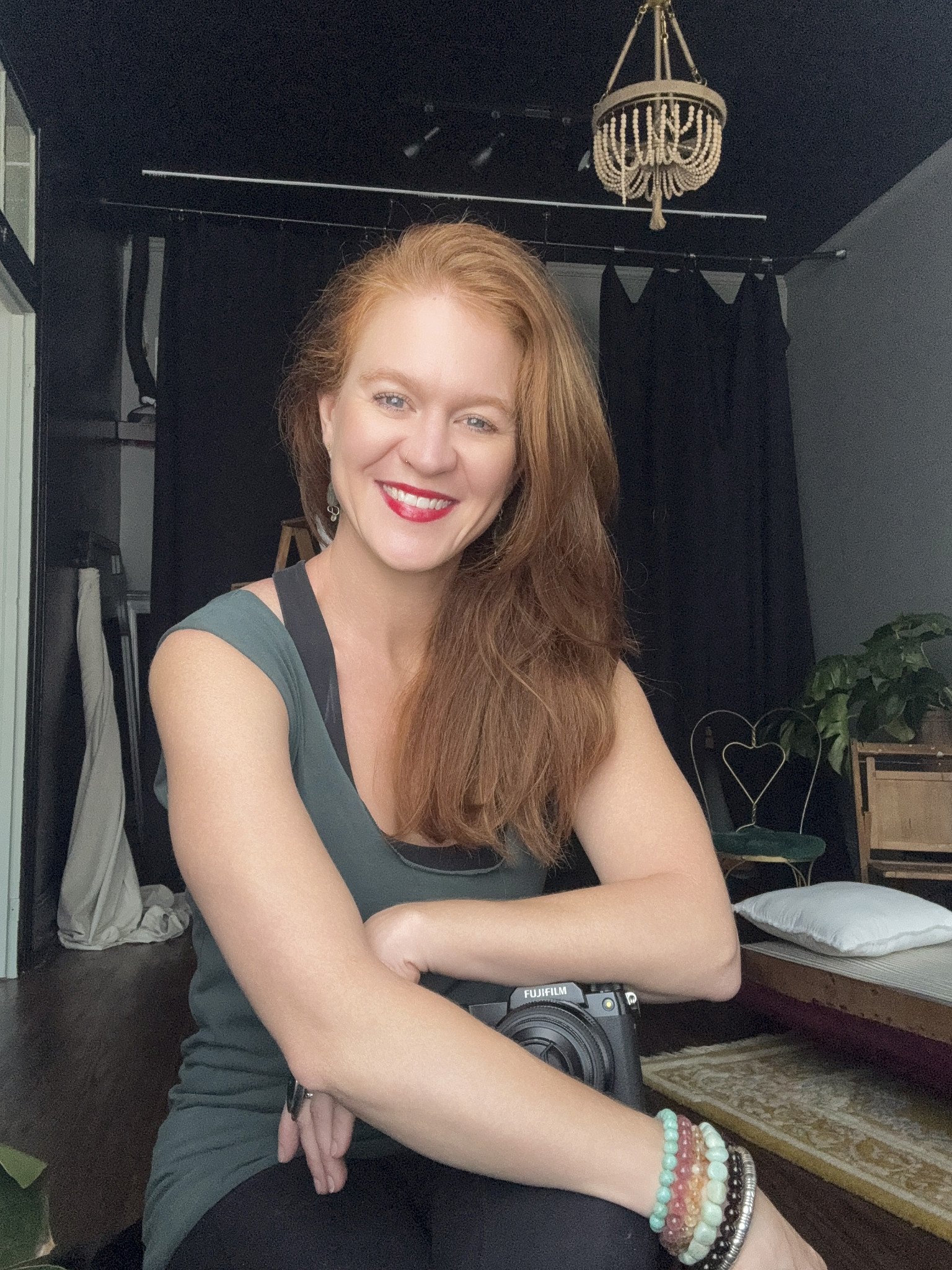 A woman with long red hair smiling, wearing a dark green top and multiple colorful bracelets, sitting indoors with black curtains, a chandelier, and a mirror in the background.