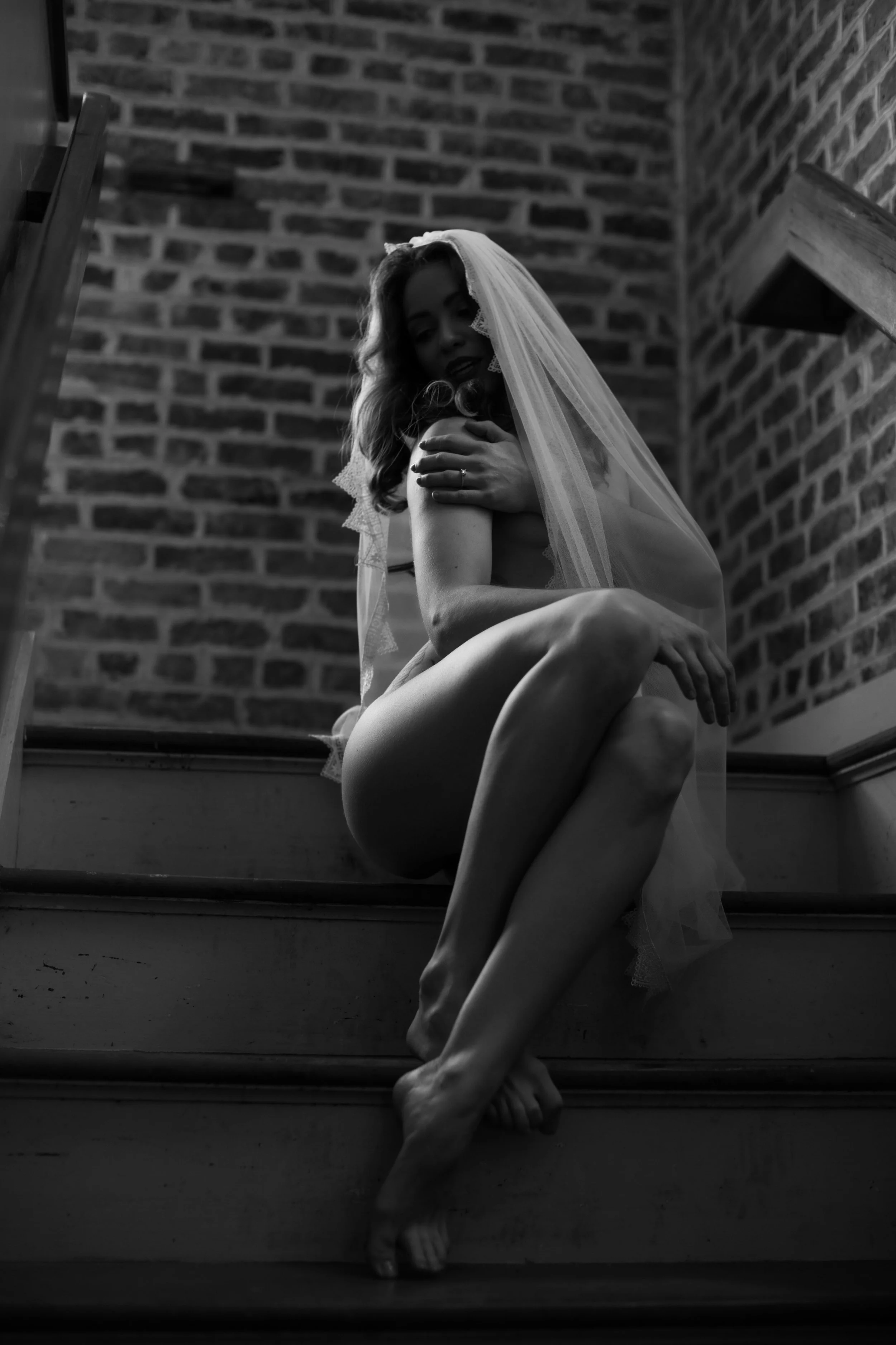 Black and white photograph of a woman sitting on stairs with a veil over her head, posing against a brick wall.