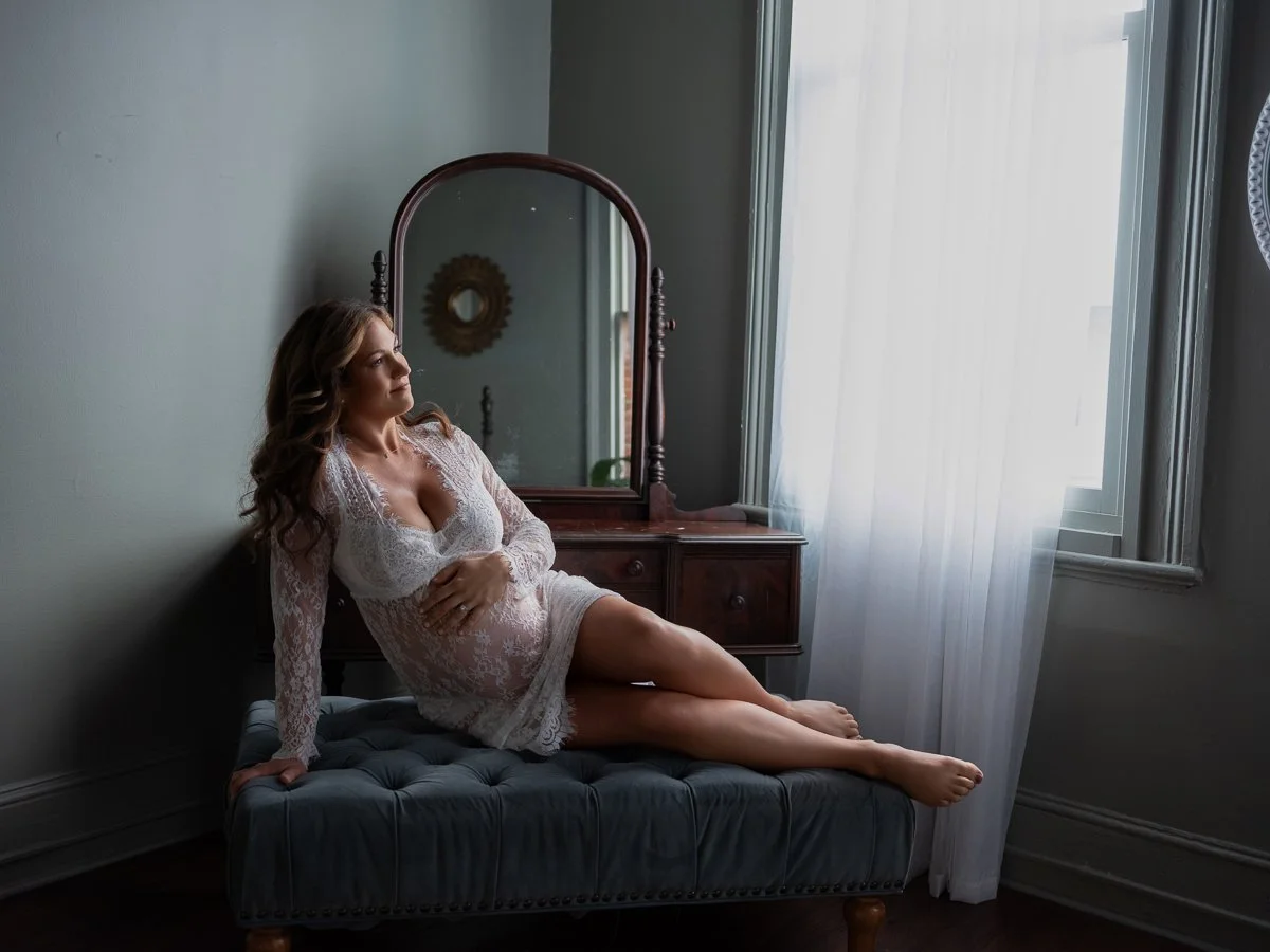 A graceful maternity boudoir portrait of a pregnant woman in white lace seated near a sunlit window, photographed by Emily Scott in Beaufort, SC.