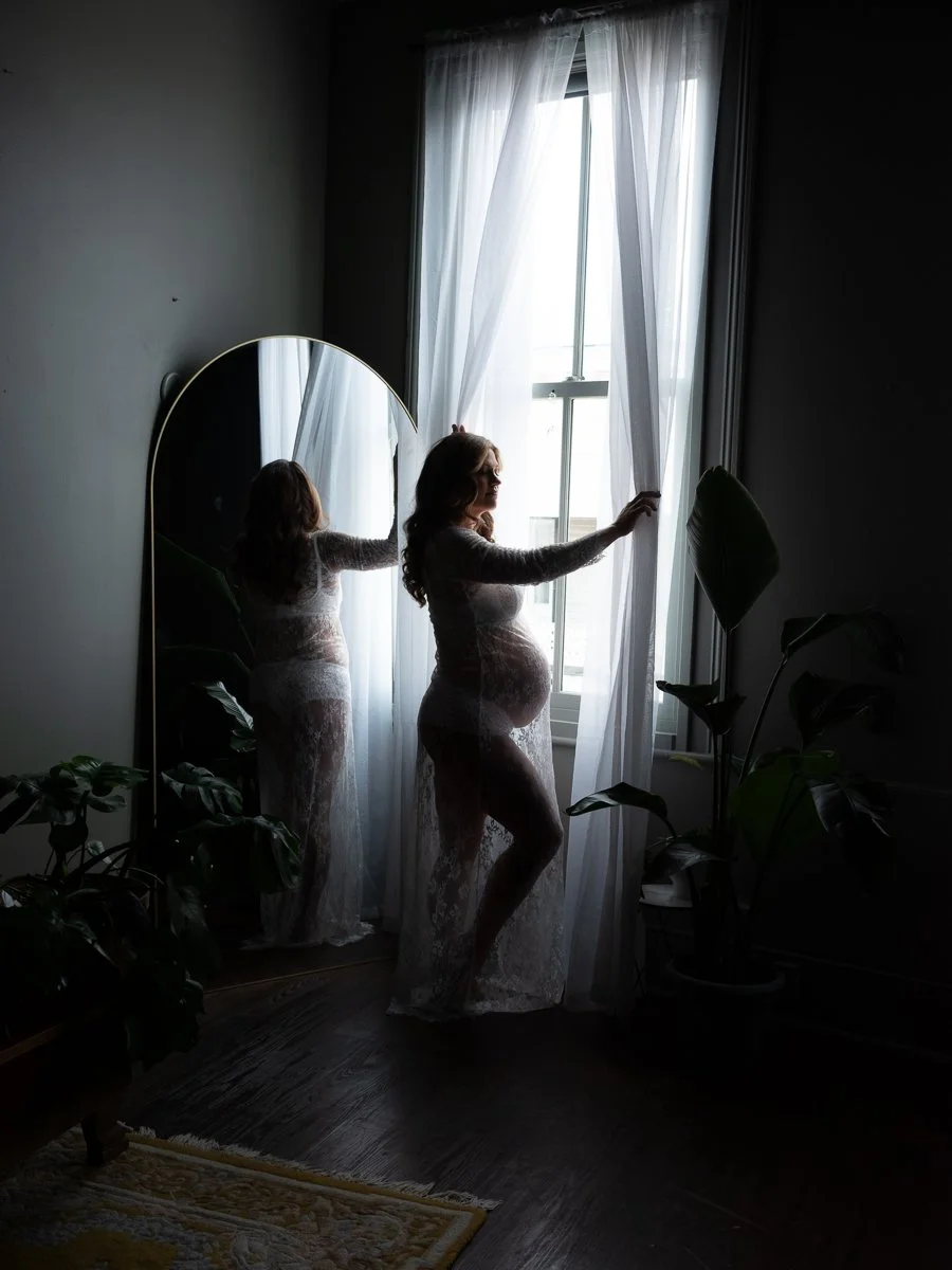 A striking silhouetted maternity boudoir portrait of a pregnant woman in a sheer lace gown at a sunlit window, photographed by Emily Scott in Beaufort, SC.