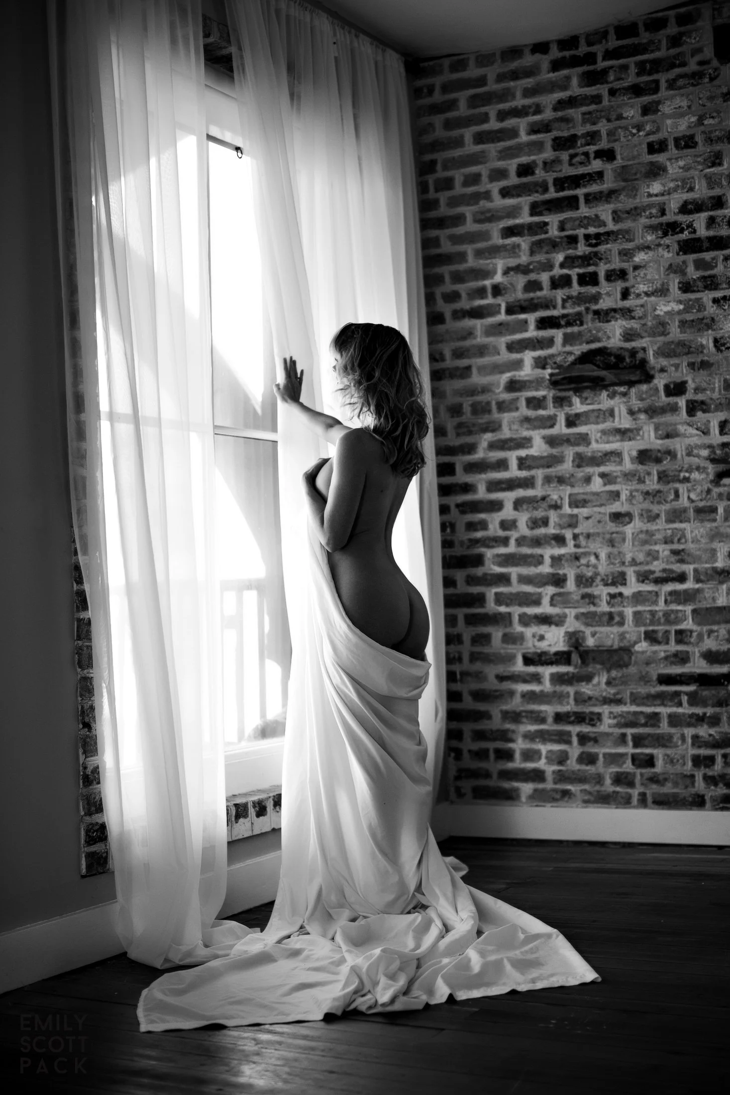 A soft black and white fine art nude portrait of a woman standing at a sunlit window near exposed brick, photographed by Emily Scott at Bay Street Studios in Beaufort, SC.