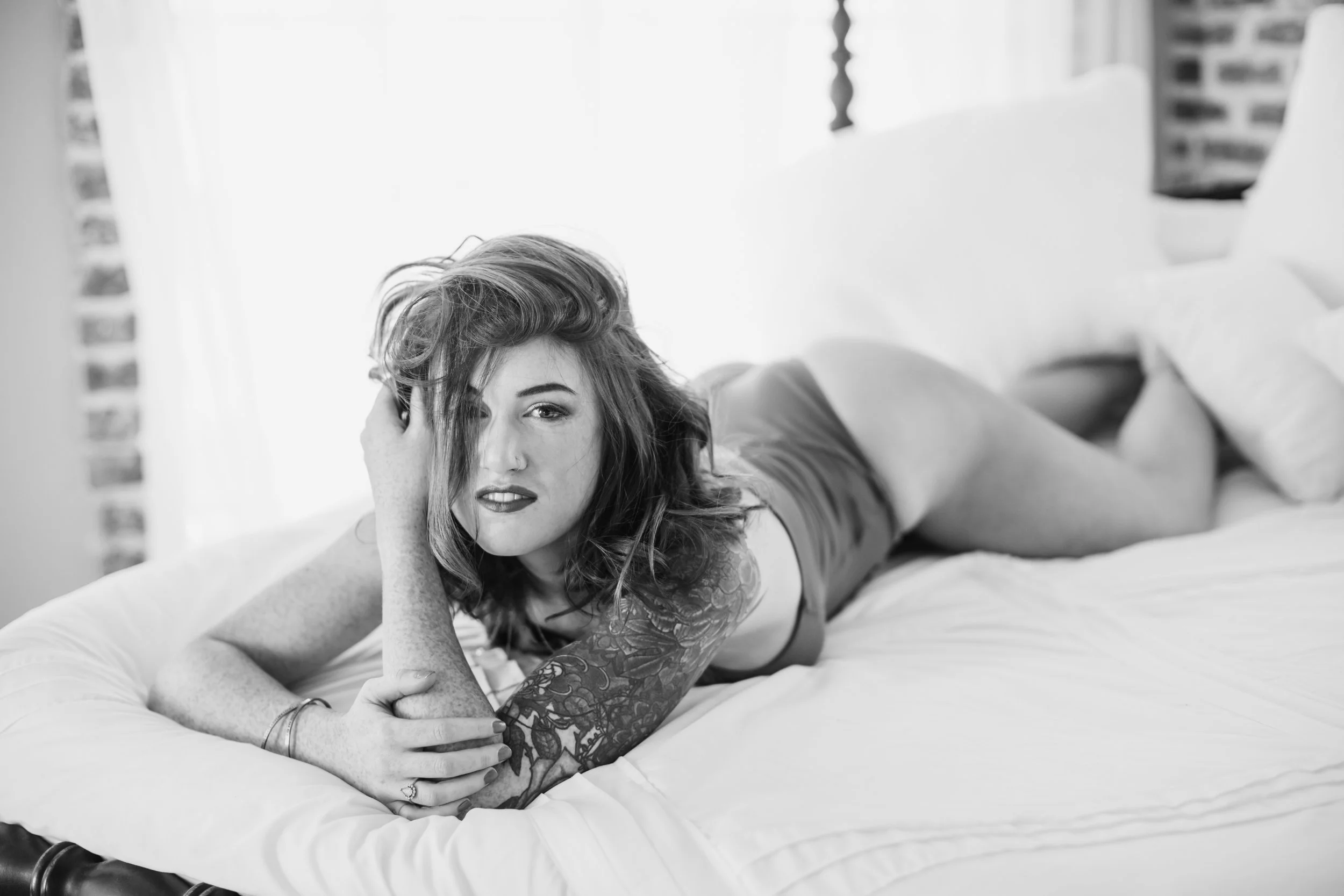 A woman with wavy hair lies on a bed, propped on her side, resting her head on her hand, in a black and white photograph.