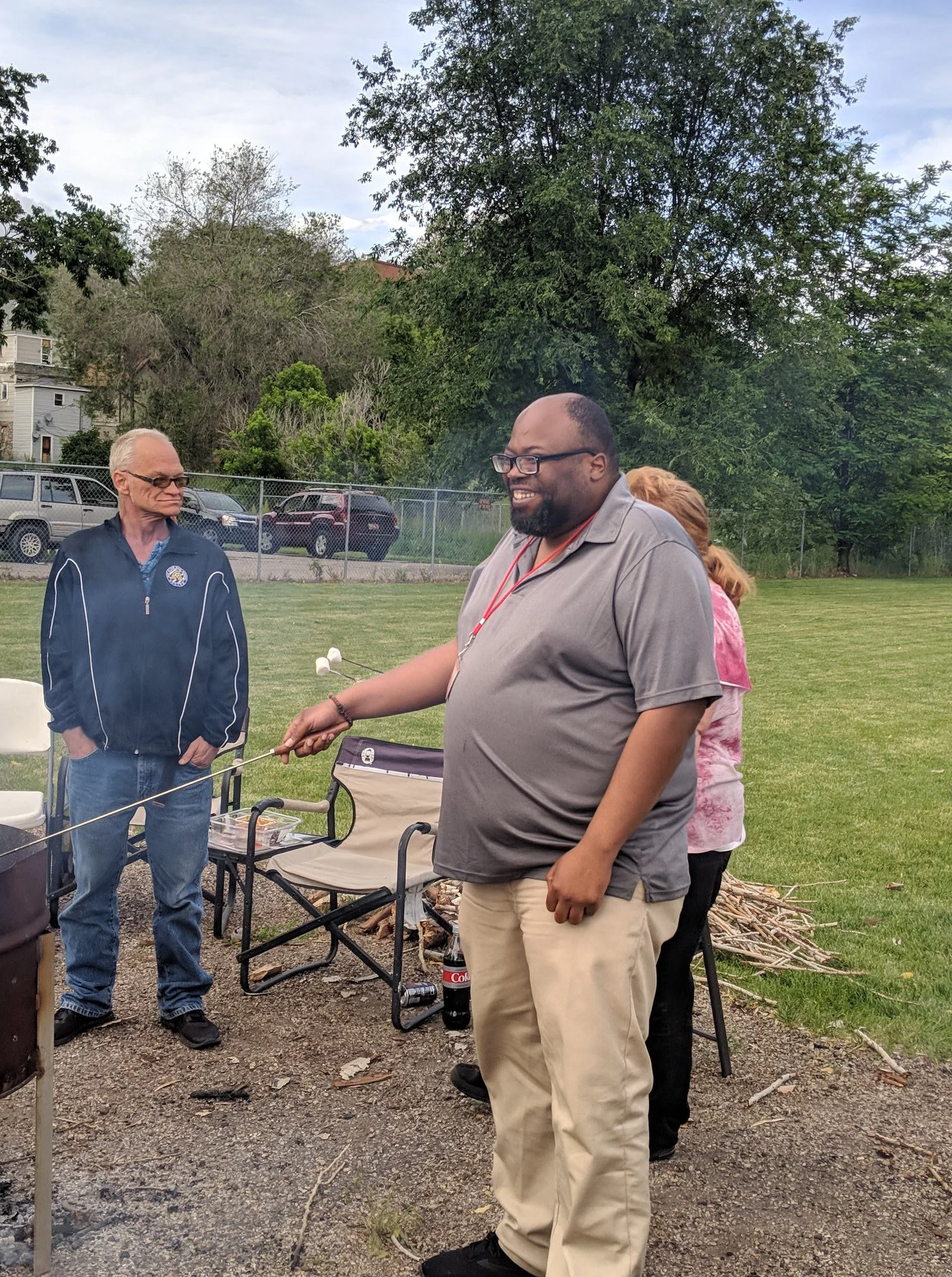 Man smiling while roasting marshmallows over a fire at an outdoor gathering with other people, chairs, trees, and parked cars in the background.