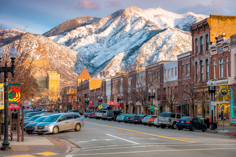 A small downtown street with parked cars, historic buildings, and mountain range with snow in the background.