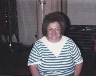 A woman with curly hair smiling, wearing a blue and white striped shirt, standing indoors near stacked chairs.