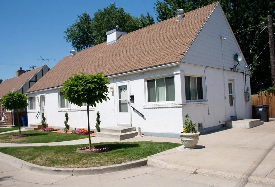 White house with brown roof, small front yard with green grass, trees, and flower bed. Sidewalk and driveway in front.