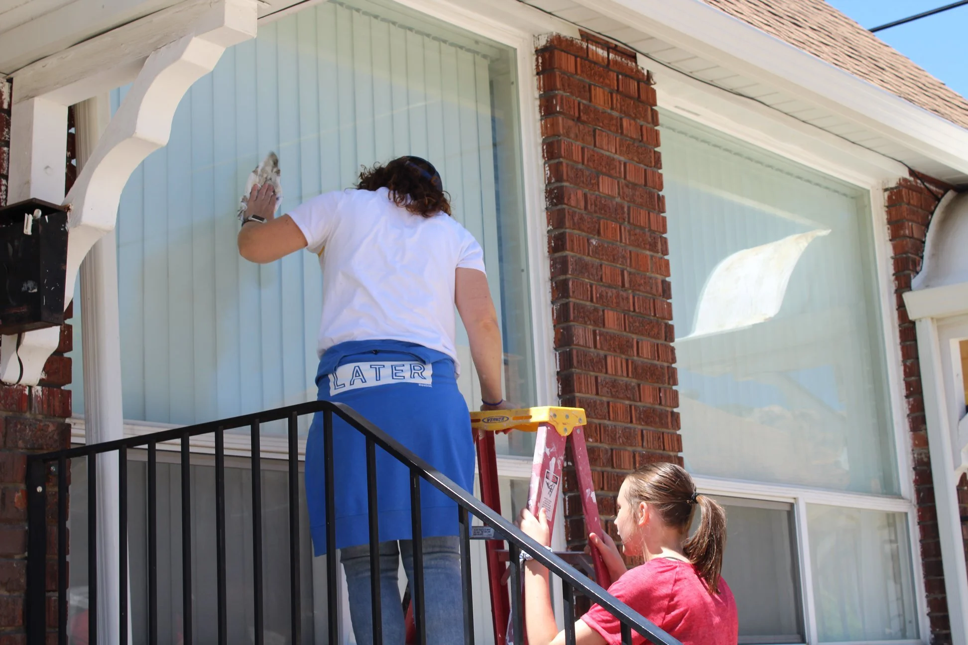 Two people standing on a staircase outside a house, cleaning the large windows. One stands on a ladder, and the other is supporting or assisting.