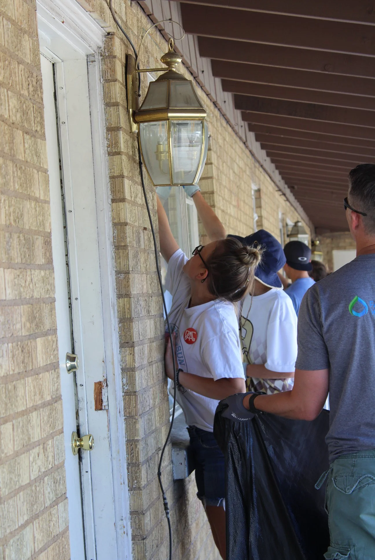 People working outdoors on a brick house wall, installing or repairing a large outdoor light fixture.