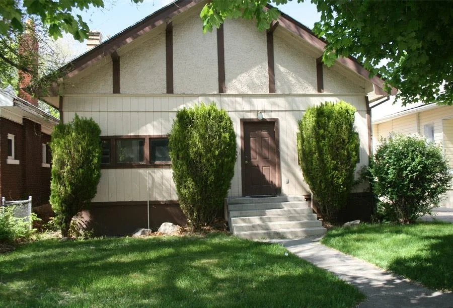 Front view of a house with a small set of stairs leading to a brown door, flanked by two large bushes, with a well-maintained lawn and trees providing shade.