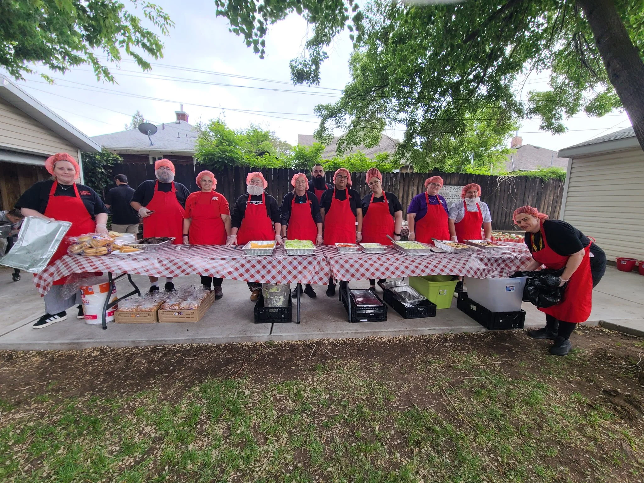 Group of ten people standing behind a long outdoor table with food, all wearing red aprons, hairnets, and gloves, in a backyard with trees and houses in the background.
