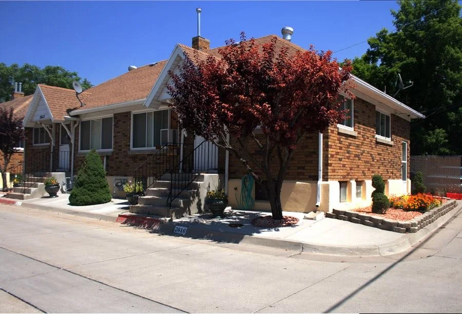 A brick residential house with a red tile roof, front steps, and a landscaped yard with trees and flowers.