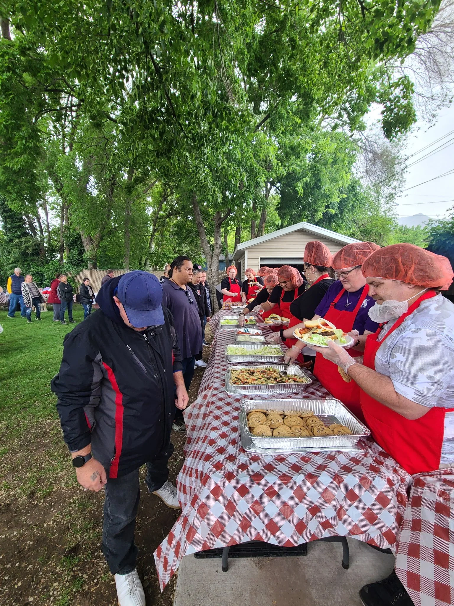 People standing in line at a buffet table outdoors, selecting food at a picnic or community gathering, with a group of people in line and others waiting in the background, under trees.