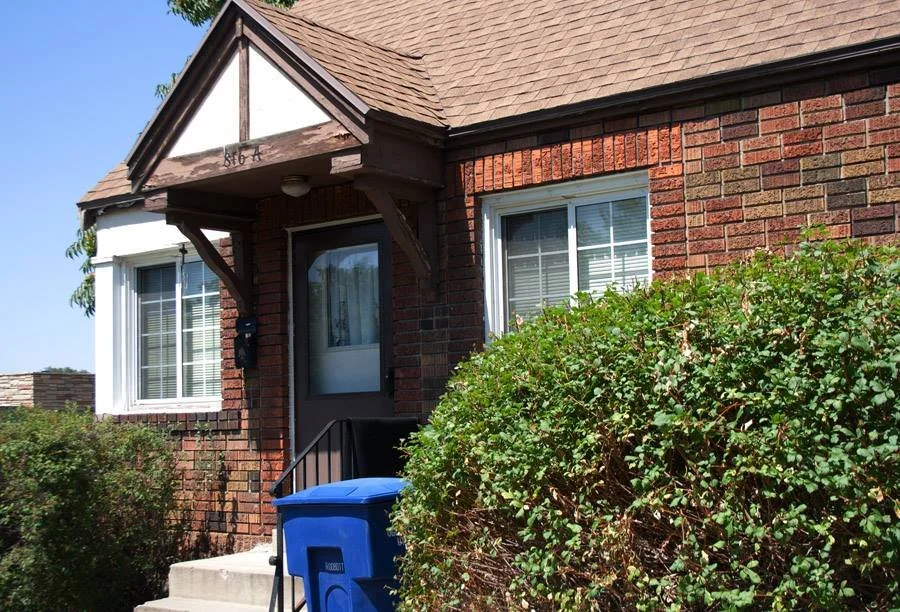 A brick house with a small porch, two windows with white blinds, and a brown door. There is a blue trash can and bushes in front of the house.
