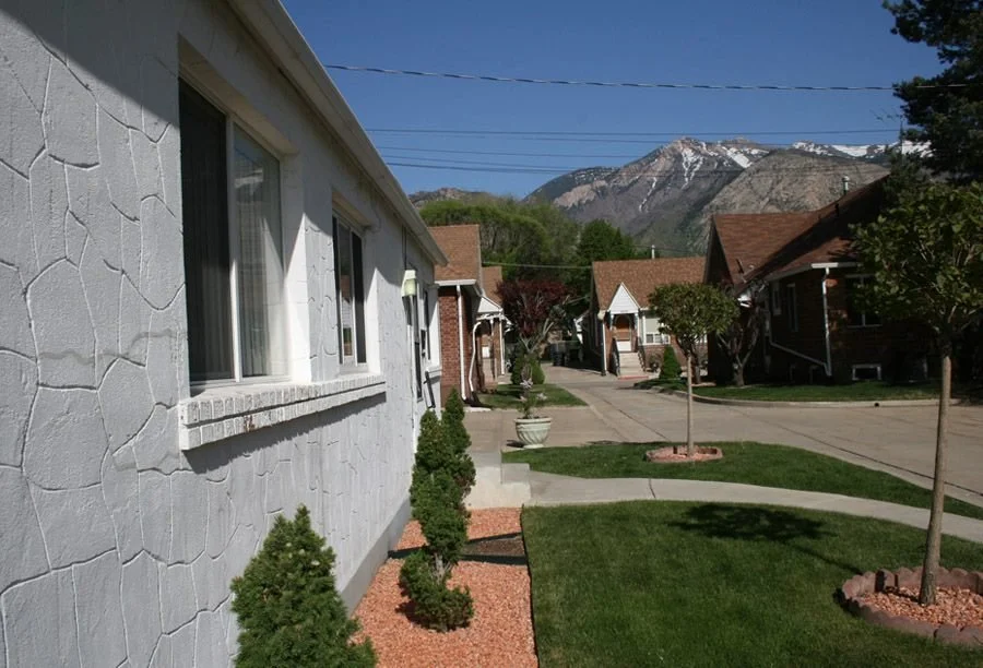 A residential street with houses, small trees, and greenery, mountain range in the background, clear blue sky.
