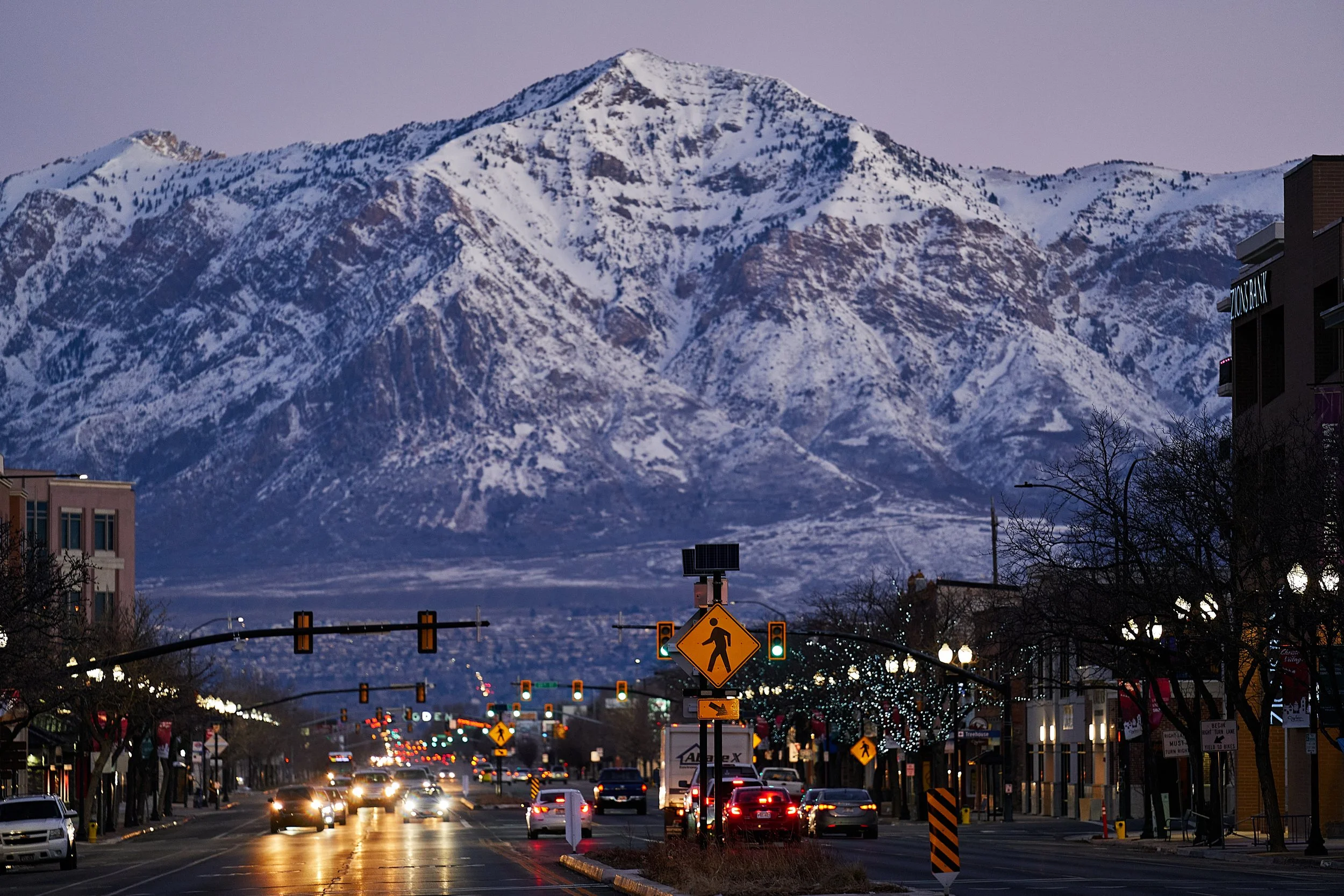 A street scene with cars driving during dusk, decorated with Christmas lights, with snowy mountains in the background.
