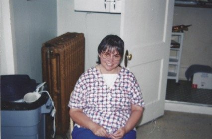 A woman with glasses and short dark hair, wearing a checkered shirt, standing inside a room near an open door.