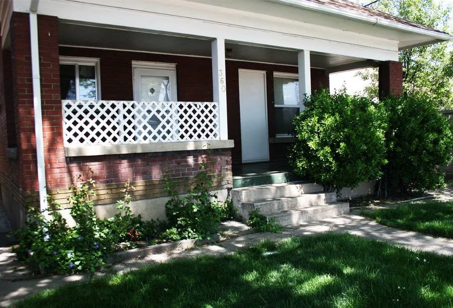 Front porch of a brick house with stairs leading up to the door, white lattice railing, green bushes, and a lawn.