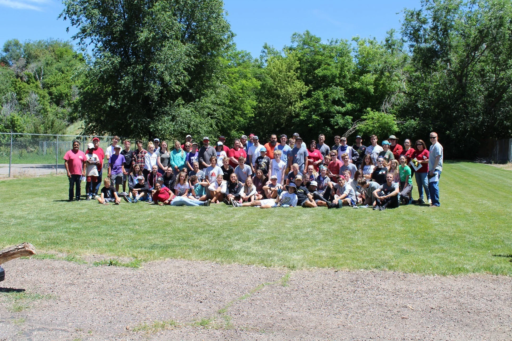 A large group of people, including children and adults, posing together outdoors on a grassy field on a sunny day with trees in the background.
