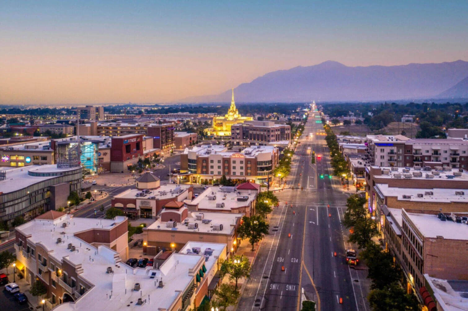 A cityscape at dusk featuring a prominent lit-up golden temple, with a long straight road leading towards it, surrounded by commercial buildings, with mountains in the background.