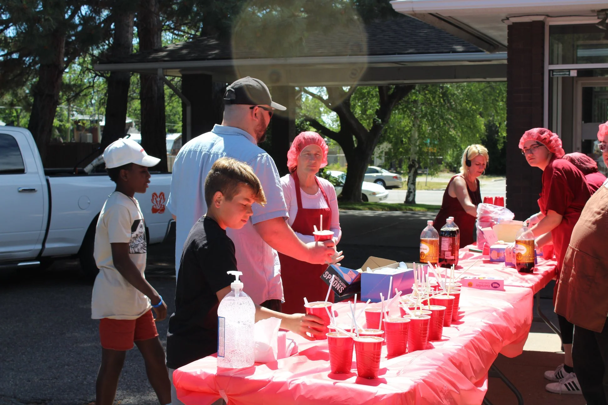 People at a community event outdoors, serving and preparing food and drinks at a long table covered with a pink tablecloth, with cups, bottles, and food items, under a shaded area.