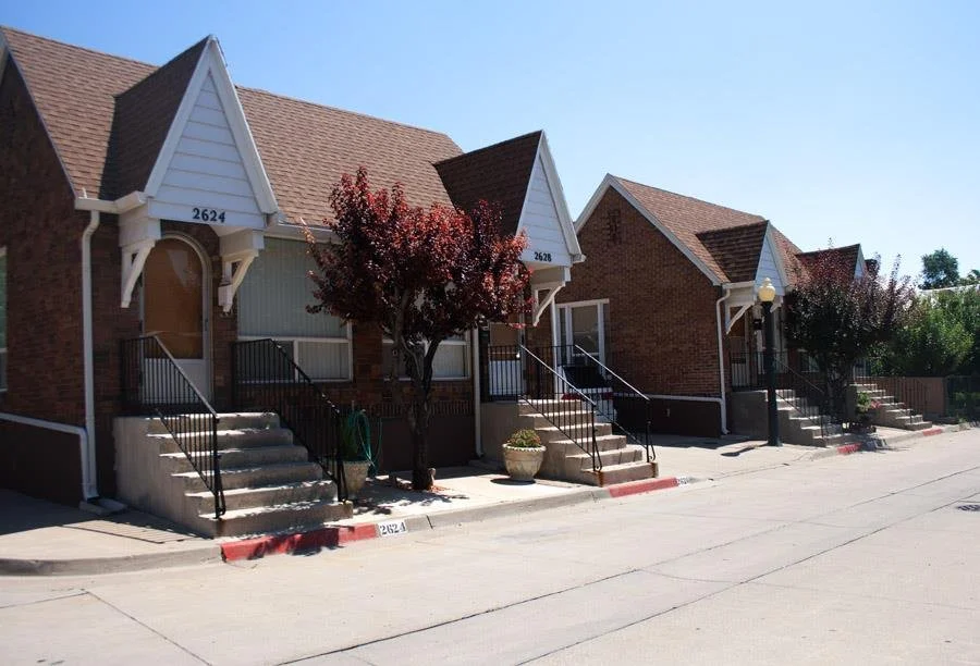 A row of three attached houses with brick facades and steeply pitched roofs, front steps, and small front yards. Two of the houses have trees in front, and a streetlamp is visible between the middle and right houses.