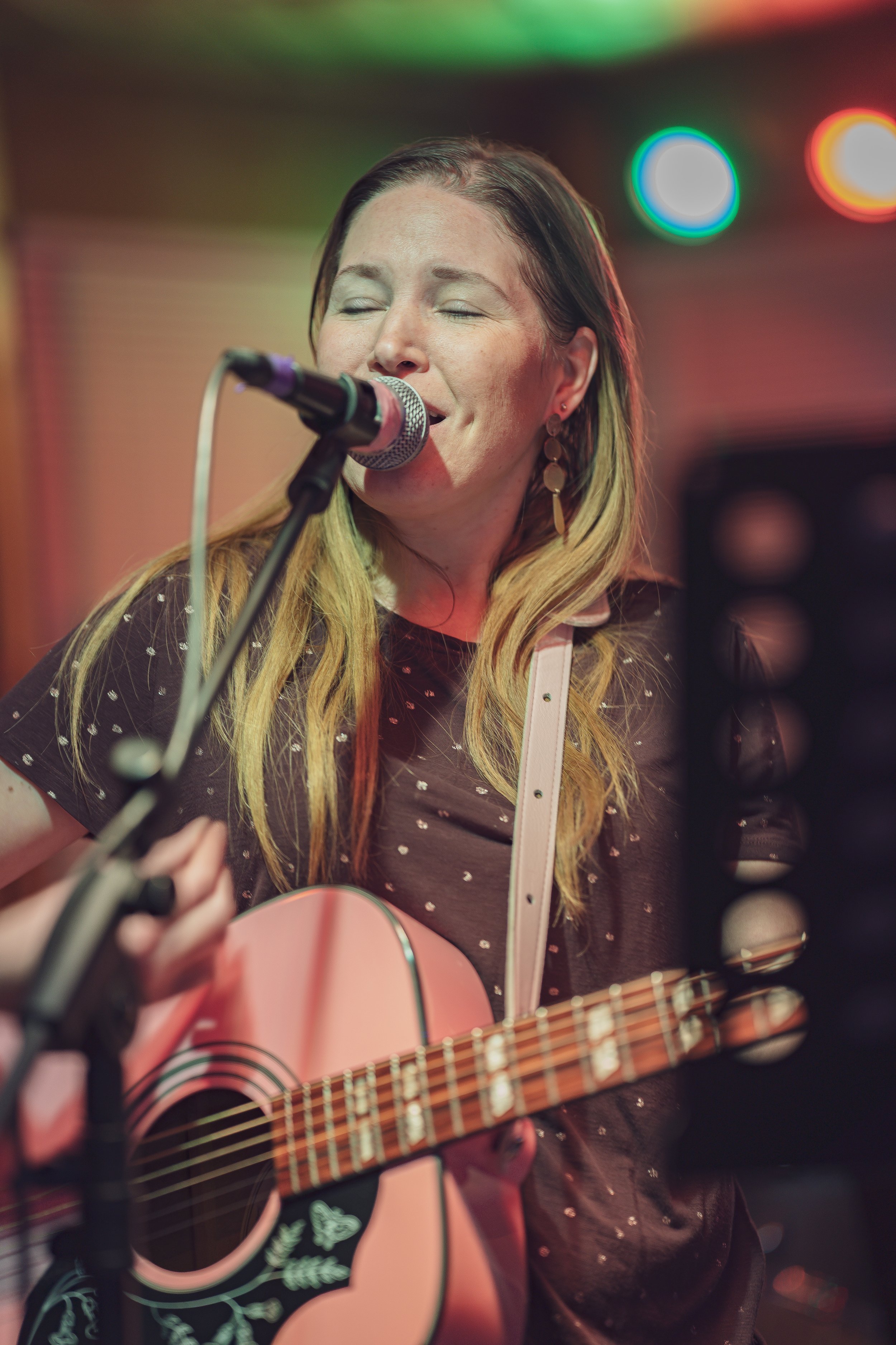 A woman with long blonde hair, eyes closed, is singing into a microphone while playing an acoustic guitar, in a setting with colorful blurred lights.