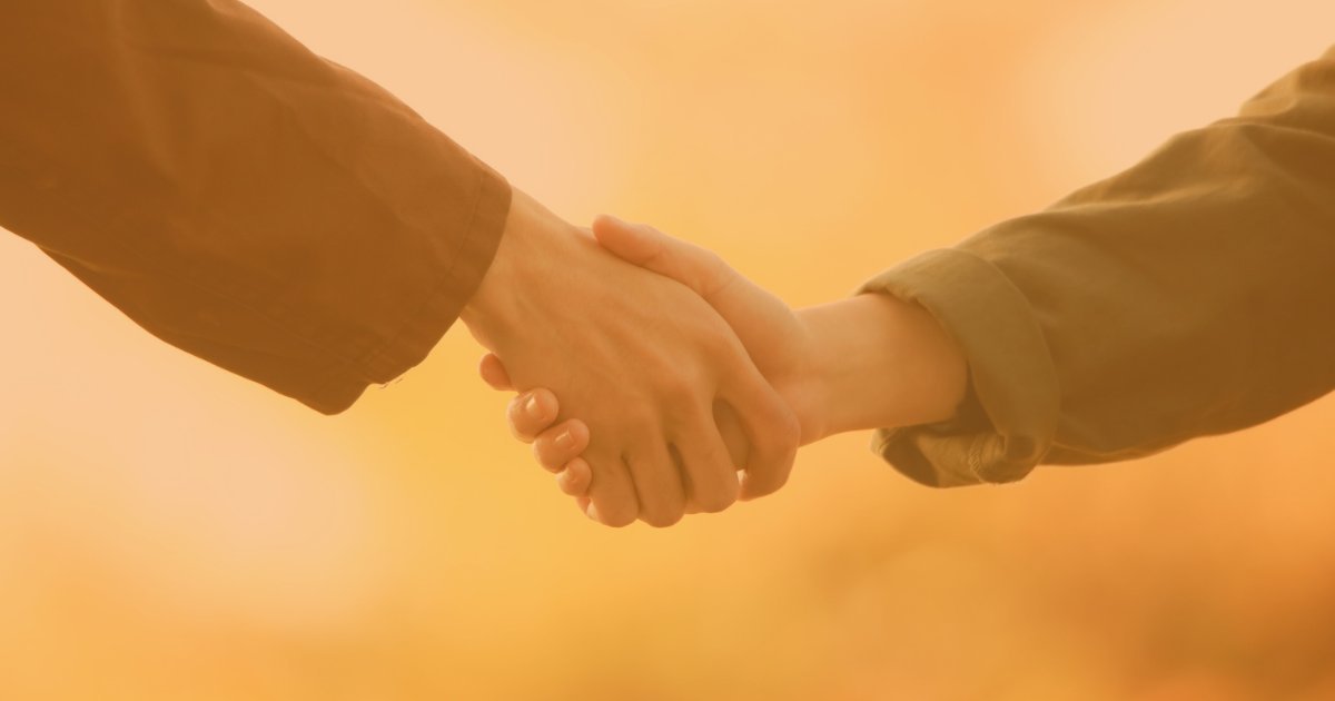 Close-up of two people shaking hands, one wearing a dark jacket and the other a brown jacket, against a yellow background.