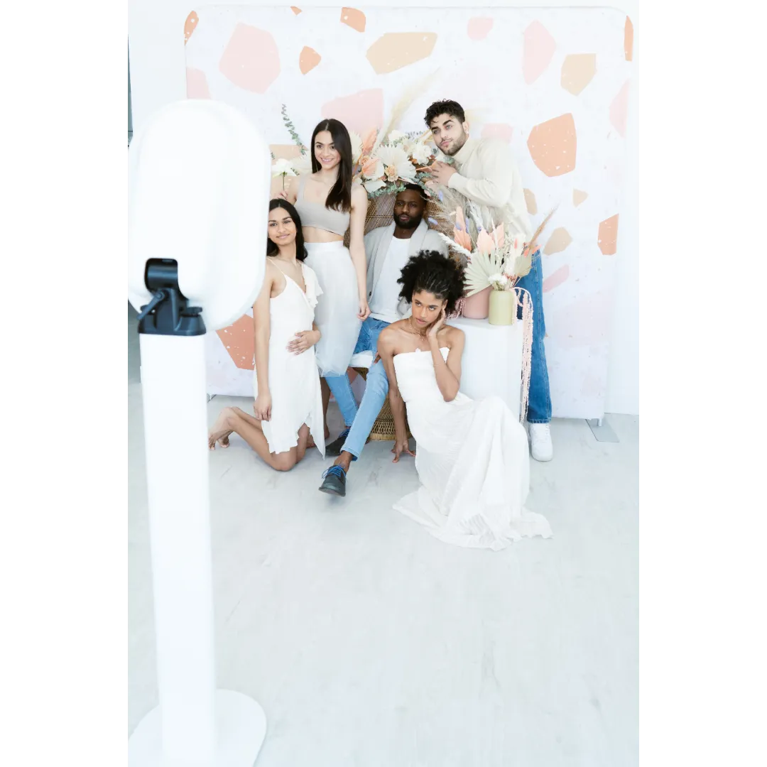 Group of six diverse young adults taking a photo in a minimalist studio with a pink and beige abstract background, decorated with white and beige flowers and dried plants.