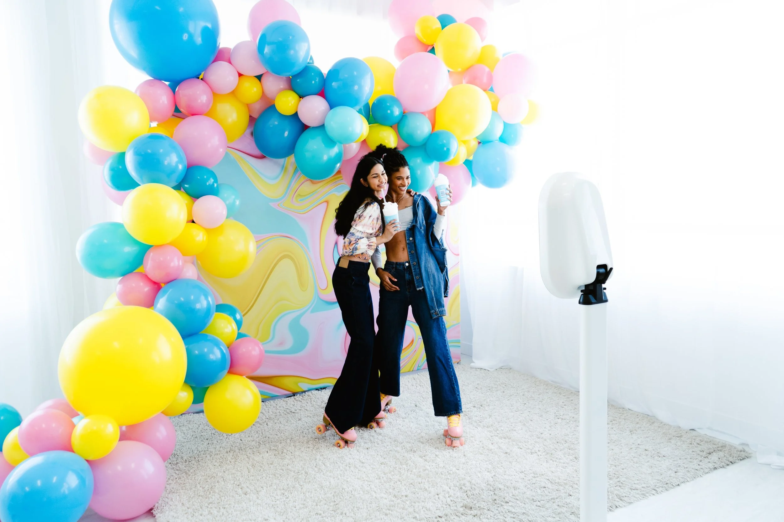 Two women roller skating and taking selfies at a colorful balloon backdrop with a marble pattern, in front of a white curtain, with a hand sanitizer station nearby.