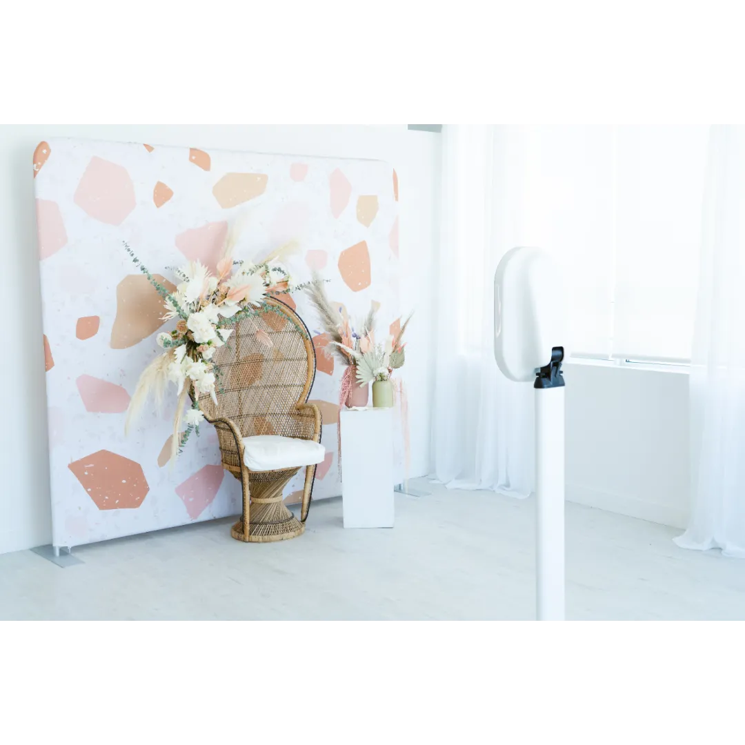 Decorative photo backdrop with a rattan chair and floral arrangements in pastel colors, set in a bright room with white curtains and a white floor.