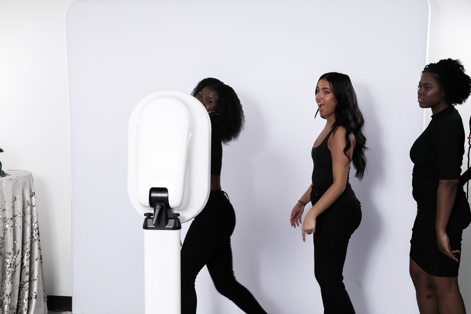 Three women standing in a line next to a white photo booth in a room with white walls.