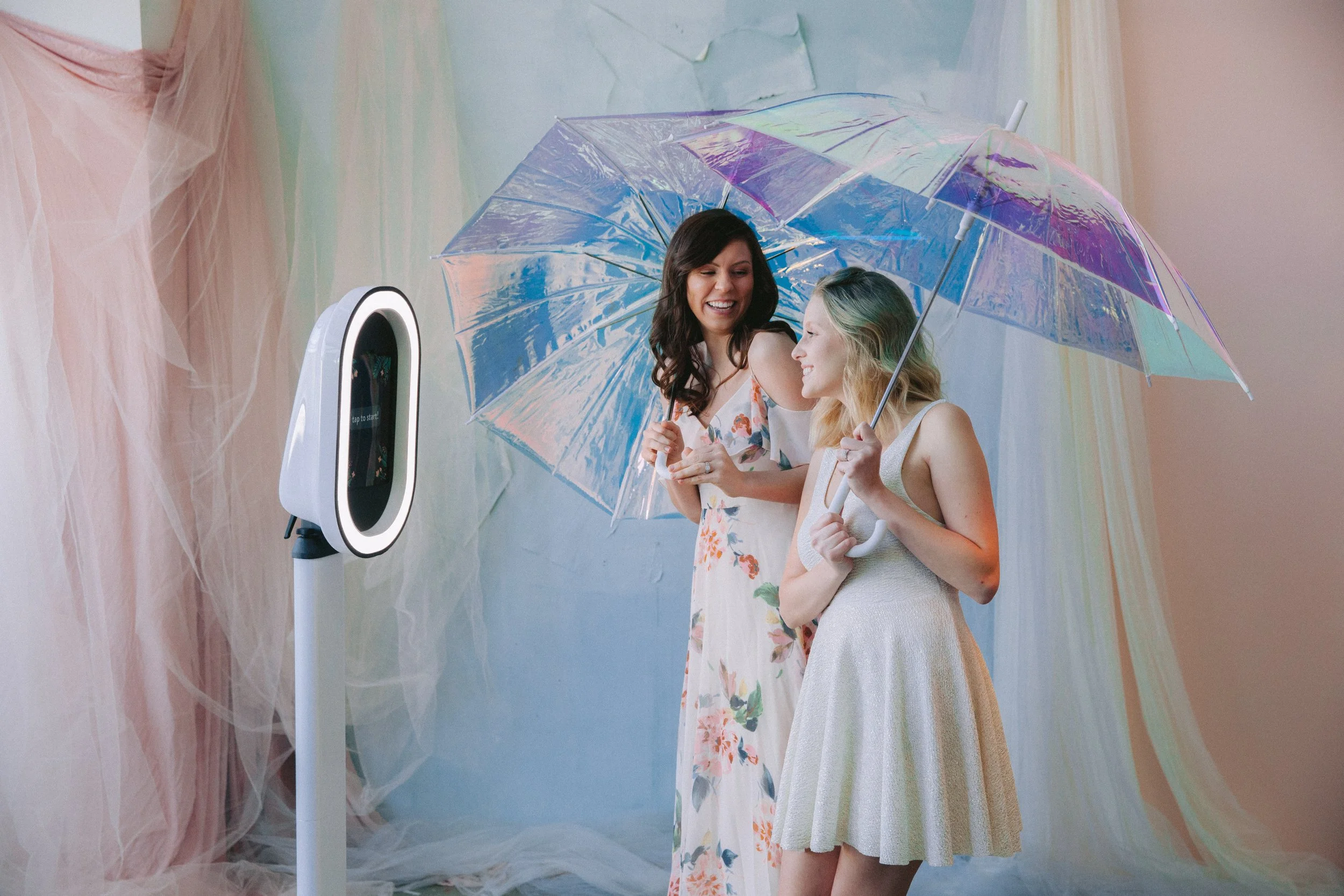 Two women stand under a colorful umbrella, smiling and interacting with a virtual photo booth.