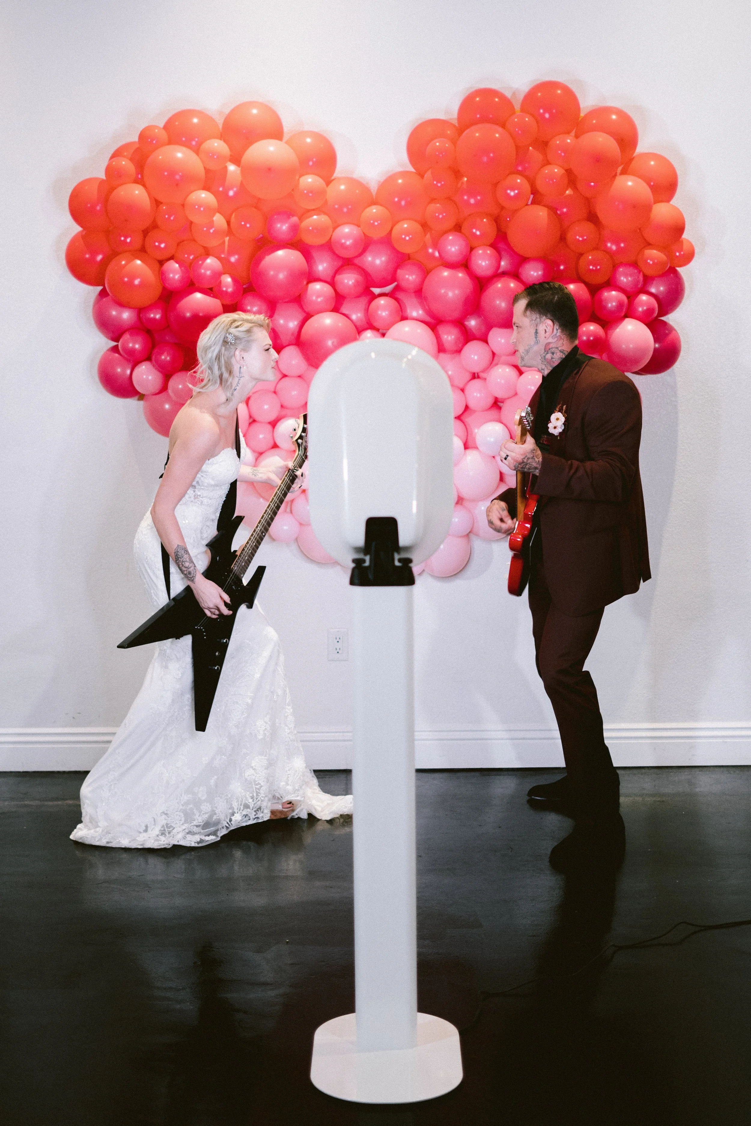 A woman in a white wedding dress and a man in a dark suit are playing guitars and facing each other in front of a heart-shaped balloon art made of pink and coral balloons. They appear to be performing or dancing in a decorated space.