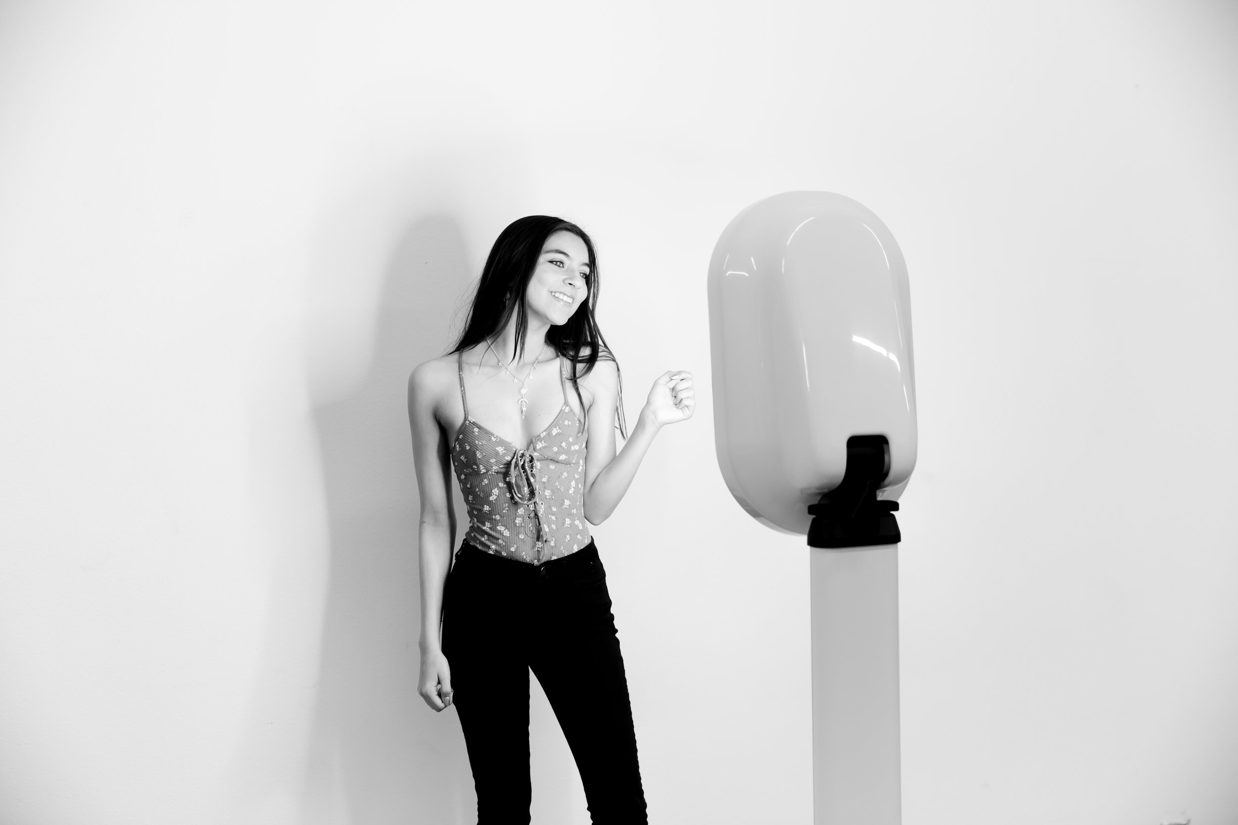 A young woman with long dark hair, wearing a spaghetti strap top and black pants, smiling and looking at a large device on a stand against a plain white wall.