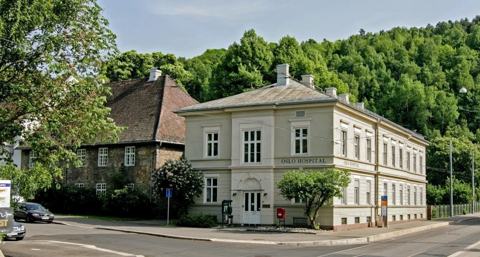 To gamle bygninger ved siden av hverandre, en av dem er Oslo Hospital, omgitt av grønne trær og bakker.