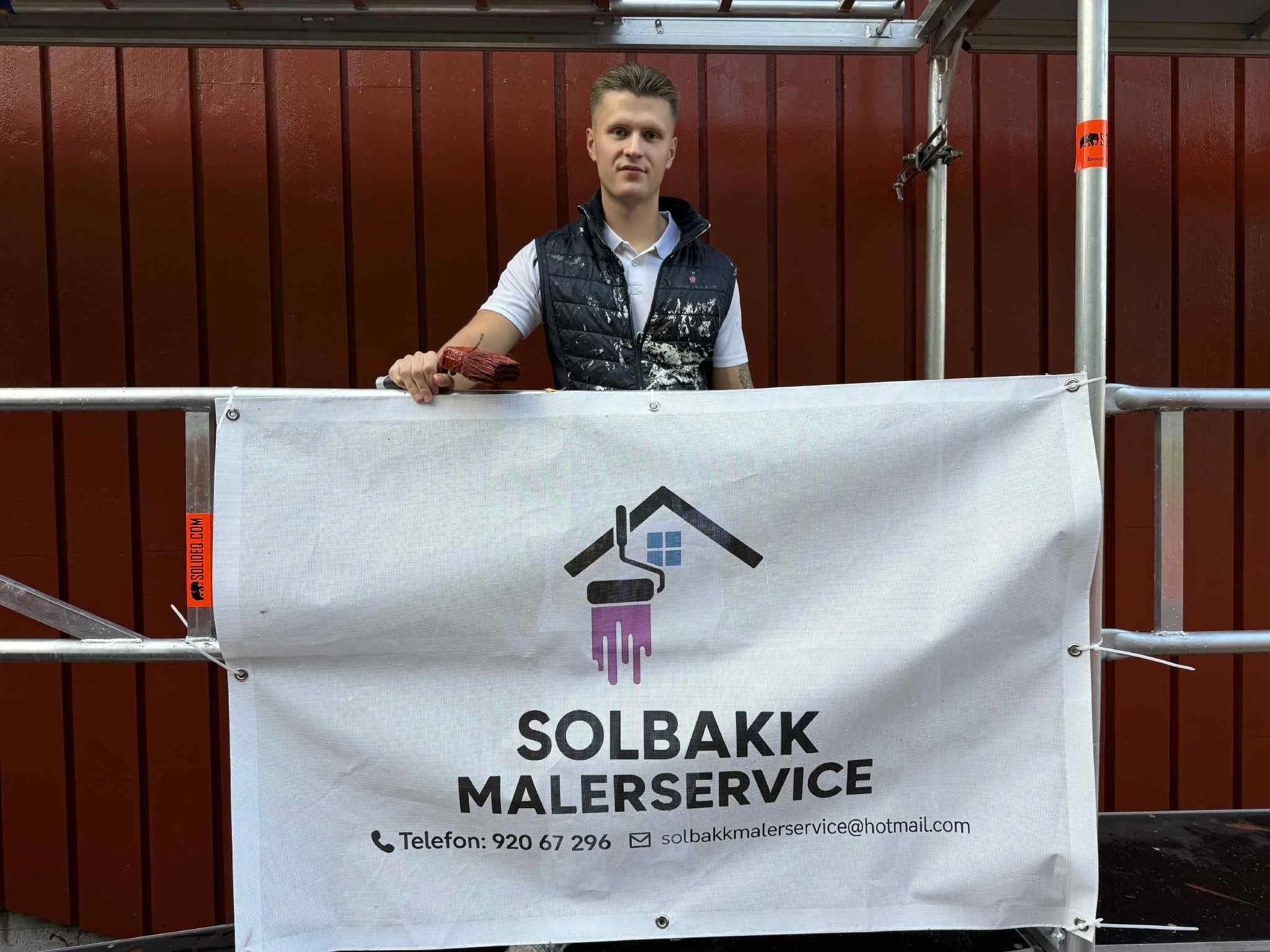Ung mann med børste som står bak en banner for Solbakk Malerservice, foran en rød vegg og stillaset.