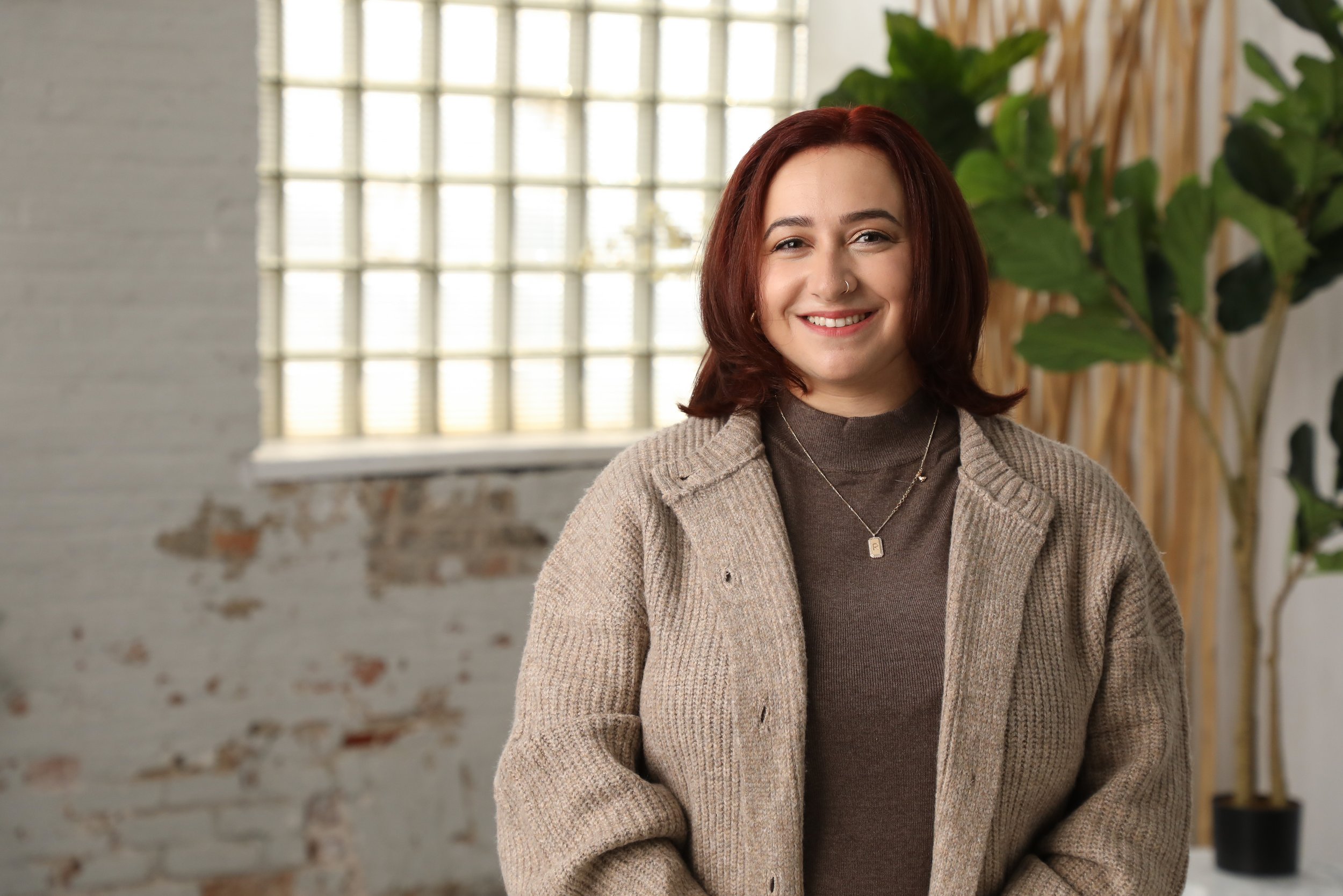 A woman with red hair smiling in a room with a large window and a potted plant.