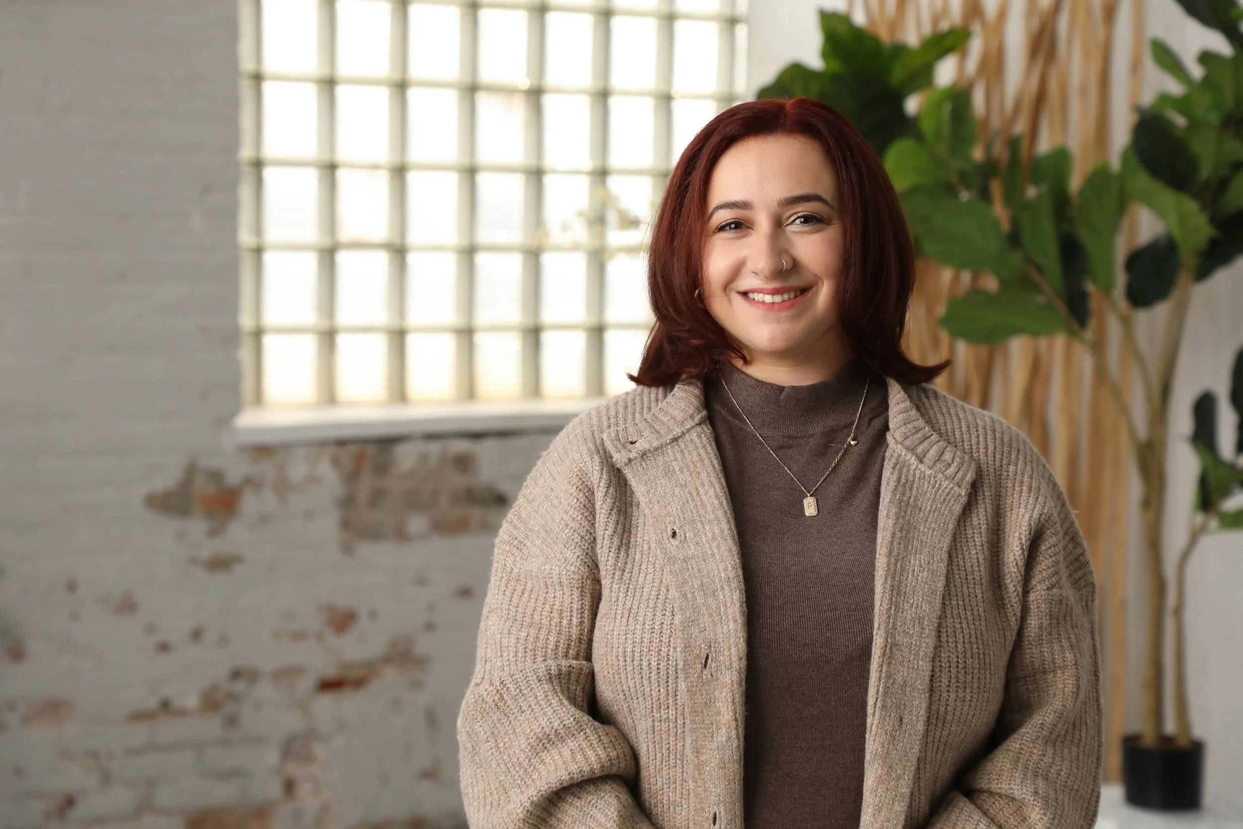 Smiling woman with shoulder-length red hair wearing a beige knitted cardigan over a brown turtleneck, standing indoors in front of a glass block window and green plant.
