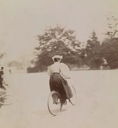 a woman wearing a hat and dressed in nineteenth century dress rides a bicycle away from the camera