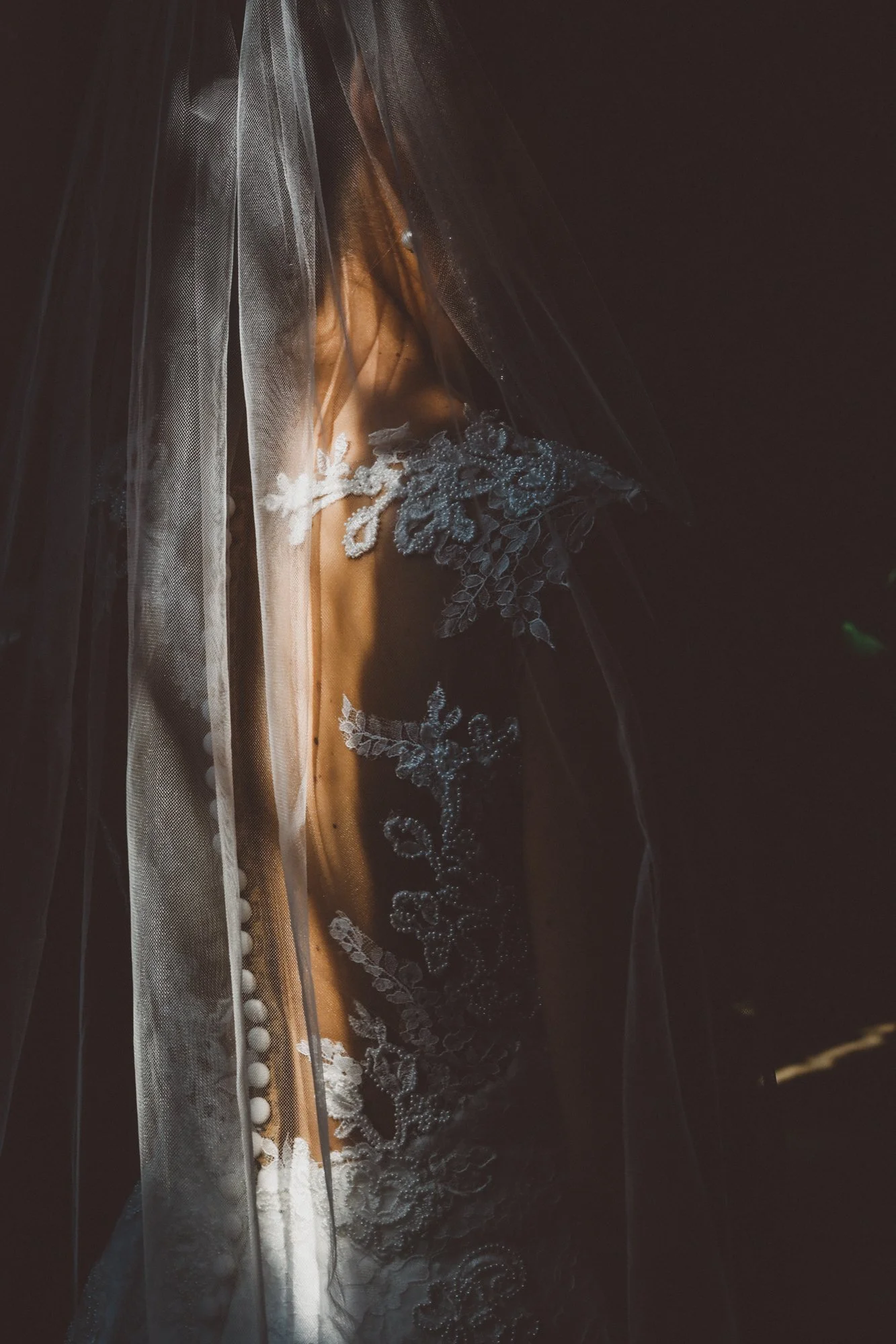 A bride wearing a wedding dress with lace and bead details, partially covered by a veil, with her face partially obscured.